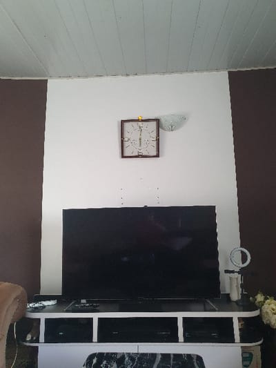 This image depicts the interior of a room, specifically a living area, located in Gero, Nigeria. The environment features a two-tone wall with a central white section flanked by vertical brown panels on the left and right. A white paneled ceiling is visible above.

Centered on the white wall, a square, dark-framed wall clock with light hands and markings is mounted. A small yellow object is positioned directly above the clock. To the right of the clock, a white, fluted wall sconce is attached. Below the clock and above the television, several small, dark circular marks are visible on the white wall.

A large, black, flat-screen television is centrally positioned on a multi-tiered TV stand. The television screen is off, displaying reflections of the room. The TV stand is composed of white and dark surfaces, featuring open compartments. Two dark electronic devices are stored within the central compartments of the stand. On the top shelf of the stand, to the left of the television, a white remote control and a black remote control are present. To the right of the television, a white ring light on a stand, a cylindrical white object, and a cluster of light-colored artificial flowers are arranged. The upholstered armrest of a light-colored furniture piece is partially visible on the far left foreground. No people are present in the scene.
