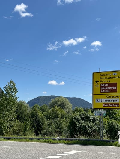 This is a roadside photograph taken on a clear, sunny day. The image features a large yellow directional sign with German text. The sign indicates directions to Salzburg, Marktschellenberg, Unterau, Roßfeld-panoramastraße, Kehlstein, and Königssee. It also points towards Berchtesgaden and "Haus der Berge." Below the main sign, a smaller, older sign displays the number 305 and an arrow pointing left. Further down, another sign indicates the distance to Salzburg (22 km) and Berchtesgaden (8.2 km), along with icons for a visitor center and a post office.
In the background, a lush green forest covers a gentle mountain slope under a bright blue sky dotted with white, fluffy clouds. Power lines are visible stretching across the upper portion of the frame. The foreground shows a portion of a paved road with white lane markings. A black pipe runs along the edge of the road, bordered by green grass and vegetation.