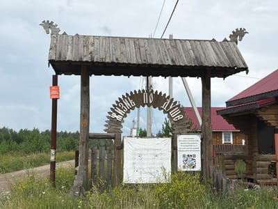 The image shows a rustic wooden archway with carved lettering in Russian that appears to say "ЖИЗНЬ ПО ДУШЕ" (Life to the soul). The archway is adorned with carved animal or mythical creature motifs on its roof. Below the arch, a weathered wooden fence is visible. A white sign hangs on the fence with Russian text and a QR code. To the right of the arch, a wooden building with a red roof is partially visible. In the background, a forest and a cloudy sky can be seen. A dirt road leads into the scene from the left. A tall, thin pole with an orange sign is located to the left of the archway. The scene is set in a rural environment, likely in the village of Kulakovo, Russia, given the provided context. The image is taken during daylight, under an overcast sky.