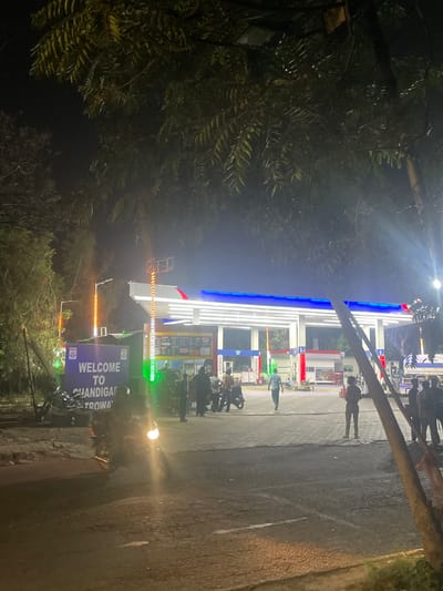 This image depicts a night scene at a gas station in Kajheri, India. A prominent sign reads "WELCOME TO CHANDIGARH TROWAY," indicating the location. The gas station is illuminated by bright lights, highlighting the canopy and the fuel pumps. Several individuals are present at the station, some standing near the pumps, others conversing. A motorcycle is visible in the foreground, with its headlight on. Trees with foliage are present in the background, and the ground is paved with brick and asphalt. The overall atmosphere is that of a busy, operational gas station at night.