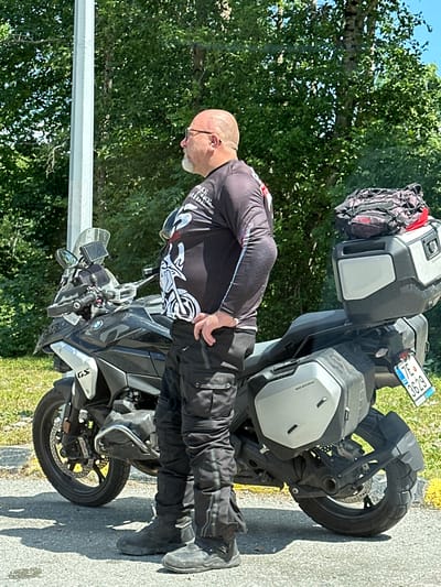 A middle-aged, bald, Caucasian male with glasses and a goatee is standing next to a black BMW GS motorcycle. He is wearing a black long-sleeved shirt with a white graphic design and black riding pants with white accents. The motorcycle is equipped with saddlebags and a top case, and a red and black bag is strapped to the top of the top case. The motorcycle has a license plate that reads "7E 3629". The man is looking to his right, with his hands on his hips. The motorcycle is parked on a paved surface next to a grassy area. In the background, there are trees and a light pole. The scene appears to be outdoors on a sunny day. The location is identified as Crkvine, Montenegro.