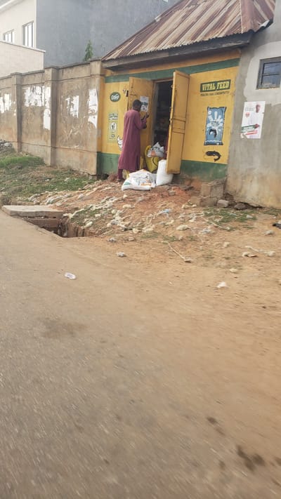 The image depicts a street scene in Kawo, Nigeria. A yellow building with a rusty corrugated metal roof serves as a retail outlet for animal feed, indicated by the "VITAL FEED" sign on the front. A person in a maroon robe stands in the doorway, possibly affixing a poster. Several sacks are visible nearby. The ground is a mix of dirt, vegetation, and what appears to be scattered waste. A paved road occupies the right side of the frame. In the distance, a residential building can be seen.