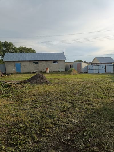 The image depicts a rural scene in Brodokalmak, Russia, on a partly cloudy day. In the foreground, a large mound of dark soil sits on a patch of unkempt grass. To the left of the soil mound, a pile of wooden planks and debris is visible. Behind the soil mound, a long, rectangular building with a corrugated metal roof and grey concrete walls stands. This building has a bright blue door and two windows. Further to the right, a smaller, shed-like metal structure with a similar corrugated roof and a pinkish door is situated. Beyond these structures, a large metal gate separates the property from the surrounding area. In the distance, a white house with a light-colored roof and trees are visible. Power lines stretch across the sky. The overall environment suggests a utilitarian or agricultural property.