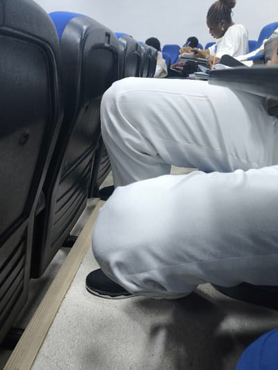 The image depicts an interior view of a lecture hall or similar academic venue located in Kızılbaş, Northern Cyprus / Cyprus. The perspective is low and forward-facing. In the foreground, the lower legs of an individual wearing white, textured trousers and black shoes with white soles are visible, bent at the knees, with feet resting on a light-colored floor. A light-toned floor strip runs horizontally in front of the seats. To the immediate left, the dark-colored, ribbed back of a lecture seat is prominent. Further into the scene, a row of blue-seated auditorium chairs extends. In the mid-ground, a woman with dark hair styled in an updo, dressed in a light-colored long-sleeved garment, is seated and actively writing in a notebook with a pen. Several documents or a folder are present on the desk surface before her. Additional seated figures are partially visible in the distant background. The room features light-colored walls and appears evenly illuminated.
