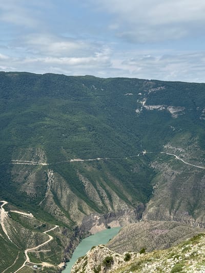 This is a high-angle, wide shot of a vast, verdant mountain range with a winding river running through a deep canyon. The mountains are densely covered with dark green foliage, with lighter green patches and rocky outcrops visible. Several unpaved roads snake along the steep mountain slopes, disappearing into the landscape. A turquoise river flows through the bottom of the canyon, with sheer, rocky cliffs on its right bank. The sky above is a pale blue with scattered white clouds. In the foreground, a rocky, sparsely vegetated slope leads down towards the canyon. The scene appears to be the Sulak Canyon, located near the village of Dubki in the Republic of Dagestan, Russia, known for its dramatic geological formations. There are no people or animals visible in the image.