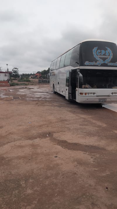 A white bus with a black top is parked on a wet, muddy, and gravelly lot. The bus has the logo "CPS COMPANY" on its back and a license plate that reads "UN 4274". The door is open and people are visible inside. The sky is overcast and grey, suggesting a rainy or humid day. In the background, there are buildings and trees, indicating an urban or semi-urban environment. The overall impression is that of a transit station or bus depot in Phao, Laos, likely during or after a rain shower.