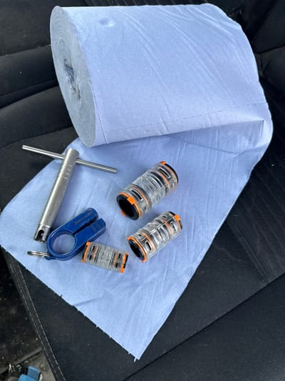 The image shows a roll of blue paper towels partially unrolled on a dark fabric surface, likely a car seat. Three clear plastic and orange mechanical components, possibly cable connectors, lie on the paper towels. A silver-colored t-handle tool and a blue plastic tool are also present. The environment appears to be indoors. No people are visible. The image provides no information that would identify the specific location as Colchester, United Kingdom.