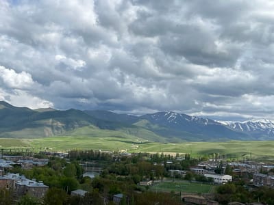 The image presents a landscape view in Sisian, Armenia, likely captured from an elevated vantage point. In the foreground, a dense collection of trees partially conceals buildings with gray rooftops, indicating a residential or urban area. A soccer field with white goalposts and visible markings is also present. A bridge spans a river or waterway winding through the town.

In the midground, rolling green hills stretch towards a mountain range. Some peaks are capped with snow, adding contrast to the scene. A road cuts through the green landscape, leading toward the mountains.

The sky is dominated by thick, gray storm clouds, diffusing the light and creating a subdued atmosphere. There are also small birds flying. The overall impression is of a scenic view within a valley, combining urban development with natural landscapes under a dramatic sky.