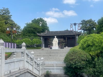 The image depicts a statue in front of a building in Hailing District, China. The statue, a light-colored figure seated in a chair, sits atop a series of concrete steps leading up to a dark-roofed building with traditional Chinese architectural elements. The building is decorated with hanging lanterns and framed by trees and greenery on either side. A stone bridge with ornate railings is in the foreground, partly obscured by foliage. The sky above is blue with scattered clouds.