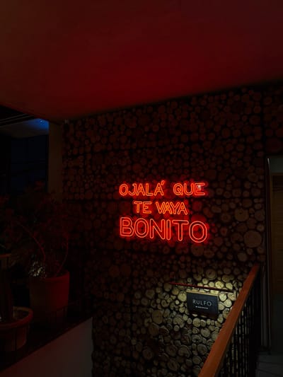 A vertically oriented photograph reveals an interior space. The walls are decorated with circular pieces of wood. A red neon sign, written in Spanish, reads "Ojalá que te vaya bonito". A rectangular sign with "Rulfo" is visible. A wooden staircase is situated near the ceiling, alongside some foliage. The overall lighting is dim, with a strong red glow.