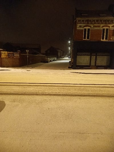 A street at night exhibits snow-covered sidewalks and a dark, empty road. Buildings line the street with visible windows and facades. Distant streetlights illuminate the way, creating a dim, long perspective. The scene evokes a quiet, cold, urban atmosphere.