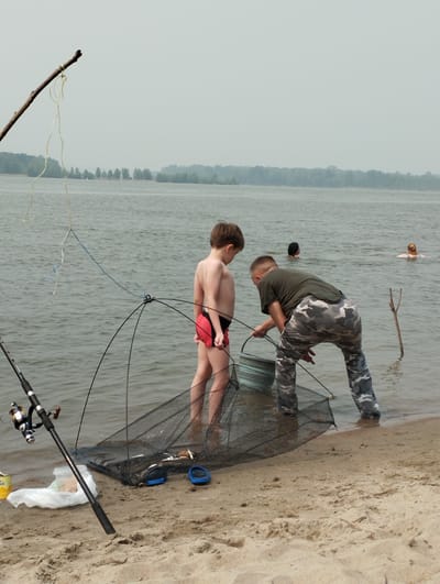 The image shows a sandy riverbank with a body of water. The sky is overcast. Two boys are in the shallow water, one wearing red shorts and the other wearing camouflage pants. They are using a fishing net and a bucket. A fishing rod is planted in the sand. Several people are swimming in the distance. The scene appears to be in Bibikha, Russia, a rural area known for its natural landscapes and recreational activities like fishing and swimming.