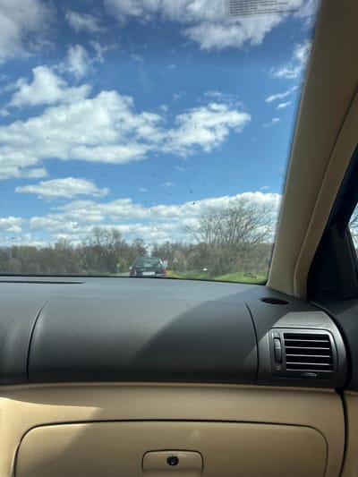 The image shows a view from inside a car, looking out through the windshield at a partly cloudy sky and trees. Another car is visible in the distance. The dashboard of the car is visible, including an air vent and glove compartment. The location is likely not Osetenovo, Bulgaria as it is impossible to determine the location from the image.
