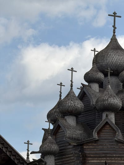The image shows the wooden domes and crosses of a traditional Russian Orthodox church, likely in the city of Nevskii Parkleskhoz, Russia. The architecture is characterized by its intricate wooden construction, with numerous onion-shaped domes covered in wooden shingles. Several crosses, some with multiple horizontal beams, top the domes. The sky above is partly cloudy with a mix of blue sky and white clouds. The church appears to be made of dark wood logs. The image is a close-up view focusing on the upper portions of the church's structure. There are no people or animals visible in the frame.