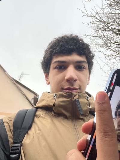 A male individual, characterized by dark curly hair and a neutral expression, is depicted in a close-up outdoor photograph taken in Meistratzheim, France. He wears a tan hooded jacket with a black backpack strap visible on his left shoulder. The individual is holding a smartphone in his right hand; the phone's screen is partially visible, displaying a mirrored image of his face, indicative of a selfie being captured. His right thumb is positioned over the lower-right portion of the phone screen. The background features an overcast sky, with a portion of a light-colored building, likely a house, to the left. This building has a sloped roof and a television antenna mounted on it. To the right, bare tree branches are visible against the sky.