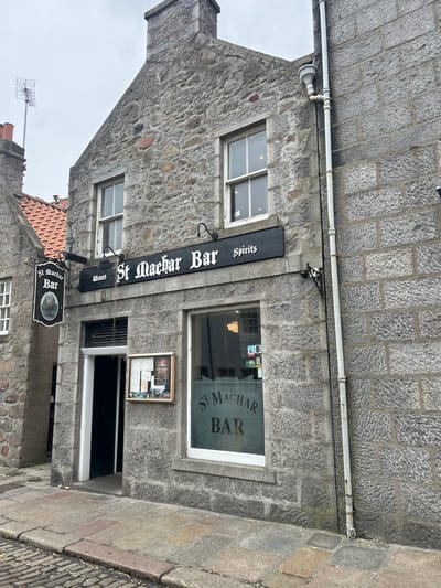 The image shows the exterior of the St Machar Bar in Aberdeen City, United Kingdom. The building is constructed of gray stone blocks, with a pitched roof section visible on the left side. Above the entrance, a black rectangular sign displays "St Machar Bar" vertically. Below the sign, two small signs that read "Wines" and "Spirits", respectively, are visible. A black circular sign with "St Machar Bar" is suspended below the Wines sign. The entrance is recessed, with a window to the right showing the name of the bar. To the right of the door and beneath the window is a framed poster or advertisement. Above the horizontal bar sign and attached to the building is a white pipe. The sky is overcast and gray. The ground slopes slightly upwards towards the right.