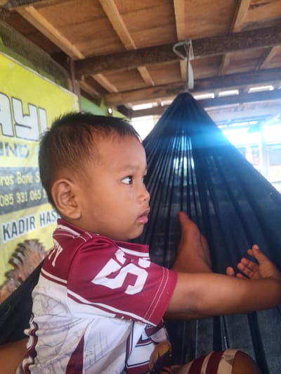 This media file shows a close-up, side profile of a young child, likely a boy, with dark hair and brown skin. He is looking to the right, with a neutral expression. The child is wearing a maroon and white striped shirt.

The background is somewhat obscured, but appears to be an outdoor or semi-outdoor setting. Above the child, a wooden ceiling or structure is visible, with beams and planks. To the left of the child, a yellow banner with text is present, though most of it is out of focus or cropped. Some visible text includes "ros Bone S", "085 331 065 1", and "KADIR HAS". To the right of the child, a dark, pleated fabric, possibly part of a hammock or mosquito net, is visible. The child's arm is resting on this fabric.

The lighting suggests it could be daytime. The scene is informal and appears to be in a casual environment, possibly a home or a local establishment. The overall impression is a candid snapshot of a child in a relaxed setting. The location context provided is Pompanua, Indonesia.