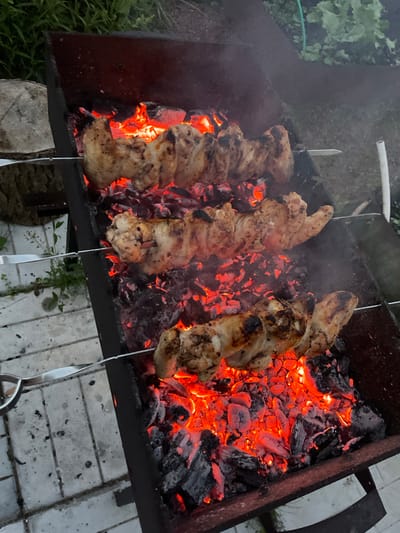 In Авиатор Снт, Russia, a close-up shot captures chicken skewers being grilled over glowing red embers in a dark metal brazier. Three skewers are visible, each holding several pieces of seasoned chicken. The meat appears to be partially cooked, with some browning and charring. Smoke rises from the coals, partially obscuring the background. The brazier rests on a paved surface made of light-colored rectangular stones. To the left, a portion of a rough, light-colored log is visible, and in the background, green foliage suggests an outdoor setting. The scene is illuminated by the intense heat and light emanating from the coals.
