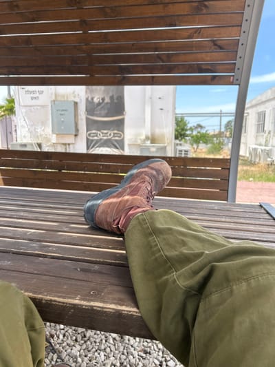 A brown leather boot rests on a wooden bench with the leg of the wearer, clad in olive green pants, resting on the bench. The bench is under a wooden pergola, and a white building is visible in the background. The setting appears to be a park or outdoor public space. The coordinates 31.173, 34.559 correspond to a location in Tze'elim, Israel. The wooden structure has Hebrew writing on it.