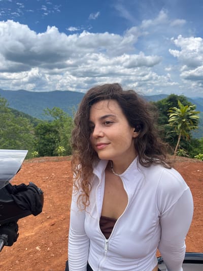 In a landscape scene in Nao Sẻ, Vietnam, a young woman with curly brown hair and fair skin is captured in a medium shot. She is wearing a white zip-up jacket over a brown top and a pearl necklace. She has black wireless earbuds in her ears and is looking to her right with a relaxed expression. Her hair is slightly windswept.
The background features a mountainous, forested landscape under a cloudy blue sky. In the foreground, red-brown soil is visible, along with a portion of a motorcycle or similar vehicle, including its black seat and a reflective windshield. The environment appears to be outdoors, likely in a rural or mountainous area of Vietnam.