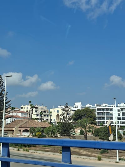 A sunlit, wide-angle view captures a coastal urban landscape in Urbanización La Cenia, Spain. The sky is a vibrant, clear blue, dotted with a few wispy white clouds. Below the sky, a row of buildings, varying in height and architectural style, forms the urban silhouette. The buildings range from mid-rise apartment blocks with multiple balconies and white facades to lower, terracotta-roofed houses. Lush green trees and palm trees are interspersed throughout the scene, adding a touch of nature to the urban setting. In the foreground, a bright blue railing runs horizontally across the bottom of the frame, suggesting the image was taken from a bridge or elevated walkway. The road and a sidewalk are visible beneath the railing, with dry grass and sparse vegetation lining the area. A street lamp stands tall on the left side of the image, and a traffic sign with yellow and white markings is visible on the right. The overall impression is one of a warm, sunny day in a Mediterranean coastal town.