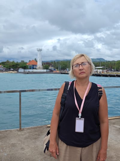 A medium shot depicts a middle-aged woman with blonde hair and glasses, standing on a concrete pier. She wears a black sleeveless top, beige pants, and carries a bag on her shoulder. A lanyard with a blank, white ID card hangs around her neck. Behind her, the turquoise ocean stretches out, with a clear view of a white lighthouse and buildings characteristic of the coastal area. The background features cloudy skies and distant hills. The scene is set in Kampung Toyapakeh, Indonesia, evidenced by the architectural style and coastal environment.