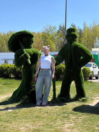 A young adult woman with tattooed arms and blonde hair in a bun is standing between two green topiary figures on a grassy lawn. The woman is wearing a white tank top, blue jeans, and tan shoes, and is holding a small pink and purple object in her right hand. One topiary figure in front of her is holding what appears to be an umbrella, and the other behind her is in a walking or striding position. Behind the figures and woman, trees, a white car, and a low-rise building are visible. The location is the city of Крошынскі сельскі Савет, Belarus. The sky is clear and blue.