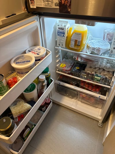 The image shows the inside of an open refrigerator, located on the floor of a kitchen in the City of Yonkers, United States of America. The refrigerator has multiple shelves containing various food items and beverages. On the top shelf, there is a carton of Kirkland Organic Milk and a jug of orange juice. Below that, there are containers of sour cream and cream cheese, along with butter, cherry tomatoes, and canned goods. Further down, there are containers of Greek yogurt, jars of pickles, jam, and barbecue sauce. There are also containers of shredded cheese and various other food items stored in plastic drawers and on shelves. The refrigerator door is open, revealing a well-stocked interior.