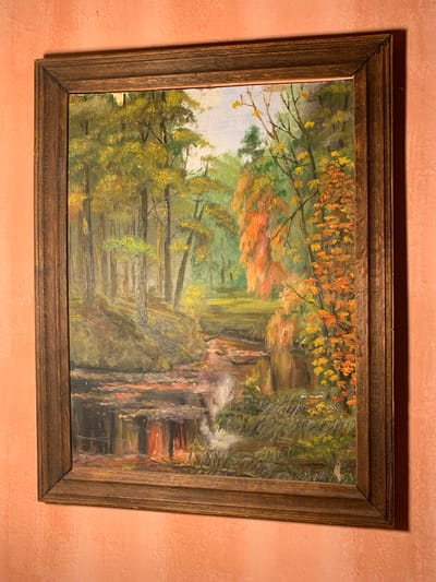 The image displays a framed oil painting of an autumn forest scene. The painting depicts a stream flowing through a wooded area with trees displaying various shades of yellow, orange, and green foliage. Sunlight filters through the trees, casting dappled shadows on the forest floor and the water's surface. The stream appears to have a small waterfall or rapid in the foreground. The painting is housed in a dark wooden frame with a textured finish. The painting is hung on a wall with a textured, peachy-pink surface. There are no people or animals visible in the painting or the scene. The location is described as Mepiskalaki, Georgia.