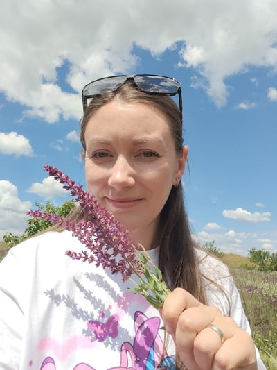 A medium shot shows a young Caucasian woman with long brown hair, wearing sunglasses pushed up on her forehead, a white t-shirt with a pink and purple graphic design, and a silver ring on her left ring finger. She is holding a small bouquet of purple flowers in her right hand, which is positioned in the foreground. The woman is outdoors in a field, likely in Novoselivka, Ukraine, as indicated by the lush green vegetation and scattered wildflowers in the background. The sky is a clear blue with scattered white clouds. The overall impression is one of a casual outdoor portrait on a sunny day.