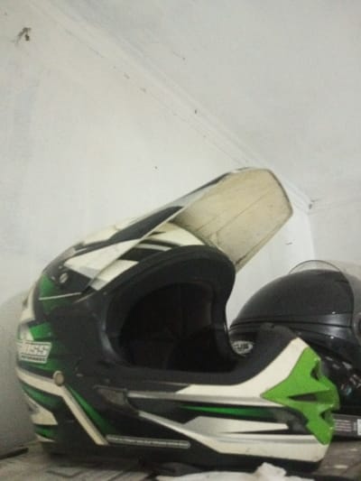 In B. Nam Ngao, Laos, two motorcycle helmets are depicted indoors. In the foreground, a full-face off-road style helmet, predominantly black with angular white and lime green graphics, rests on a light-colored surface. A "CROSS" brand logo is visible on its left side, and its upper visor is elevated. Partially obscured behind it to the right, a second, dark-colored (likely black) full-face street motorcycle helmet is visible, with its clear visor lowered. Both helmets are stationary. The background consists of a plain, light-colored wall, showing minor surface imperfections and faint horizontal lines. No individuals or active interactions are present in the scene.