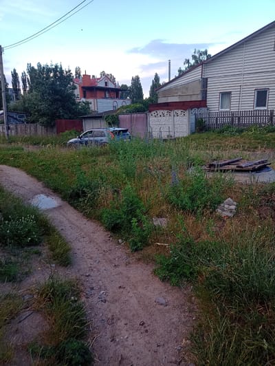 This media file depicts an outdoor scene with a dirt path winding through overgrown grass. In the background, there are several structures, including a multi-story building with light-colored siding and a dark roof, possibly a house or an apartment building. Adjacent to this structure is a wooden fence. Further in the distance, another building with red and white brickwork and a prominent orange roof is visible, with trees surrounding it. A car, partially obscured by the vegetation, is parked on the left side of the path. The sky is a light blue, suggesting daytime, and the overall impression is one of a rural or suburban setting with elements of neglect due to the overgrown grass and the presence of the abandoned car. The lighting suggests it could be late afternoon or early evening, with soft, diffused light. No people are visible in the image. There is no visible text.