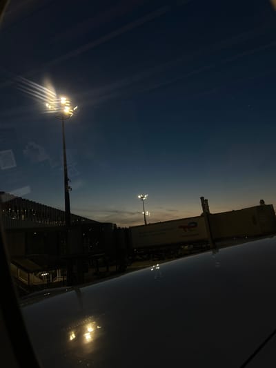 This image depicts a nighttime or twilight scene at an airport, likely Charles de Gaulle Airport (CDG) in Roissy-en-France, France. The perspective is from inside a vehicle or aircraft, as evidenced by the reflection on a dark, glossy surface in the foreground, which appears to be a window or a wing.
Dominating the upper left of the frame is a tall light pole with multiple bright, upward-facing lights, casting strong beams into the deep blue sky. The sky shows faint contrails or atmospheric disturbances. To the right, a jet bridge is visible, connected to a terminal building. The terminal has a modern architectural design with rows of illuminated windows.
On the jet bridge, the logo and text "TotalEnergies, la compagnie de toutes les énergies" and "TotalEnergies" are clearly visible, indicating a partnership or service provided by this energy company at the airport. Further to the right, another set of lights is visible on a shorter pole.
The overall impression is one of an active airport during low-light conditions, with artificial lighting illuminating the infrastructure against the darkening sky. The glossy foreground reflection adds a dynamic and slightly abstract element to the scene, obscuring details of the immediate environment below. No people are clearly discernible in the image.