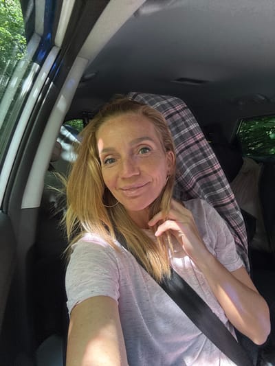 The image shows a woman with blonde hair and freckles taking a selfie inside a car. She is wearing a light pink t-shirt and a seatbelt. Behind her, a plaid blanket is visible. The car's interior is dark, with sunlight filtering through the windows, illuminating trees and foliage outside. The location is identified as Portovoe, Russia.