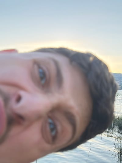 A low-angle, close-up, selfie-style photograph captures a person's face in the foreground, with a blurred background depicting a body of water and a distant hillside with buildings under a bright, pale blue sky. The person's face is tilted, and their eyes, nose, and mouth are visible, but the image is significantly out of focus. The body of water appears calm, with some reflections and reeds or grasses growing along its edge. The hillside suggests an urban or semi-urban environment. The lighting indicates it is likely daytime, possibly late afternoon or early morning, given the soft light. The image was taken outdoors. The location is the city of Emek HaYarden Regional Council, Israel.