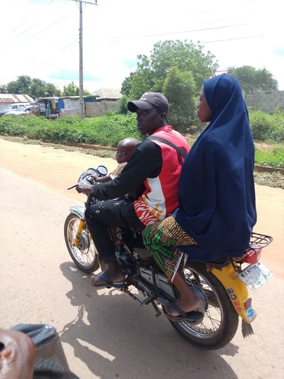 Three people are riding on a motorcycle on a dusty road. The rider is a man wearing a black baseball cap and a red vest over a black shirt, with black pants. A young child, wearing a yellow and white striped shirt, is sitting in front of him, facing forward. A woman is sitting behind the rider, wearing a navy blue hijab and a vibrantly patterned skirt. The motorcycle has a yellow rear fender with the number "2026" written on it in blue. A license plate is attached to the fender, though the text is mostly illegible. The scene appears to be in a rural or semi-urban area, with sparse vegetation and some buildings visible in the background, including a utility pole with overhead wires. The sky is bright and overcast, suggesting daylight, likely mid-morning or afternoon, with a slight haze. The overall mood is one of everyday transport.