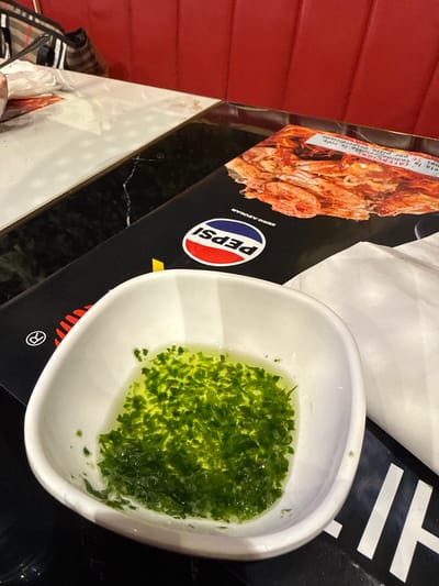 The image shows a close-up of a white bowl filled with a green liquid and finely chopped herbs, likely a sauce or dressing. The bowl is placed on a black surface with a Pepsi logo and an image of cooked shrimp on a menu or placemat. In the background, a red upholstered seat is visible, suggesting a restaurant setting. The lighting is bright, highlighting the textures and colors of the food. The environment appears to be indoors, possibly a casual dining establishment. There are no people clearly visible in the image, and no specific location in Carretera De San Martín, Spain, is identifiable.