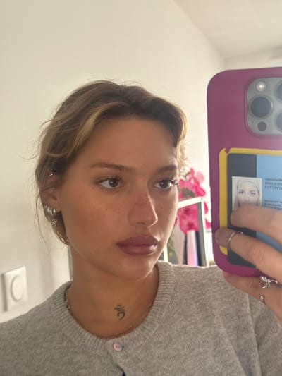 A young adult female is depicted in a close-up, self-portrait taken indoors in Nogent-sur-Marne, France. Her right hand holds a magenta-cased smartphone featuring a prominent triple-lens camera array. A yellow cardholder attached to the phone case displays a partially visible, inverted identification card with text indicating "Nogent" and a name like "Matilde" or "Mathilde." The subject has light brown hair with blonde highlights, noticeable freckles on her nose and cheeks, and brown eyes. She wears light pink lipstick. Her left ear is adorned with a helix piercing and two lobe piercings, one featuring a small hoop with a dangling element. A small, dark tattoo, resembling an Om symbol, is visible on the right side of her neck. She wears a light grey knit cardigan with a single light pink button visible and a thin, beaded necklace. Her right index finger wears a plain silver band, and her right pinky finger wears a more ornate silver ring. The background consists of a plain, light-colored wall with a white electrical outlet or switch on the left. Blurred pink floral decorations are reflected in the background behind the phone, suggesting an indoor residential environment.