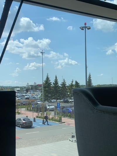 The view from a window shows a parking lot filled with cars in front of a large building, likely an airport. The sky is bright blue with white clouds. A man in a black shirt and pants is walking across a crosswalk. There are trees and a tall light pole with multiple floodlights. The view is from the interior of a building in Pyrzowice, Poland, near the Katowice Airport.