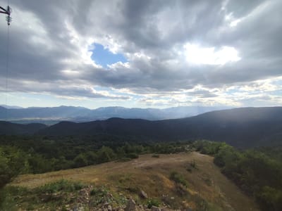 The image depicts a panoramic view of a mountainous landscape under a cloudy sky. In the foreground, a barren, rocky slope with dry grass leads to a lush green forest covering the lower slopes of the mountains. In the distance, a series of layered mountains recede into a hazy horizon. The sky is filled with dramatic clouds, with sunlight breaking through in several areas, casting rays of light. A single, isolated power line pole is visible in the upper left corner of the frame, silhouetted against the sky. The scene appears to be located in or near Gorno Dryanovo, Bulgaria, a region known for its mountainous terrain and natural beauty.