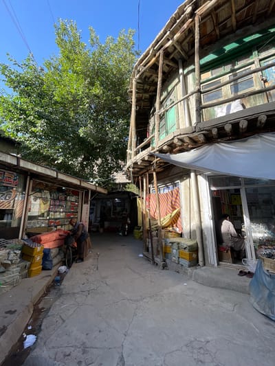 The image depicts a narrow street scene in Kabul, Afghanistan, characterized by traditional architecture and bustling market activity. On the right, a two-story building with wooden balconies and green window frames exhibits signs of age. The lower level features a shop with a glass storefront displaying various goods. A person, dressed in traditional Afghan attire, is seated inside the shop. On the left, another shop displays goods on shelves and stacked items are placed outside on the ground. A man is attending to items near a bicycle in front of this shop. The street is paved with concrete and shows some wear. A drainage channel runs along the side of the street, with some debris visible. Overhead, a large tree with green foliage provides shade, and electrical wires are strung across the sky. The overall atmosphere suggests a vibrant, everyday scene in a local market.