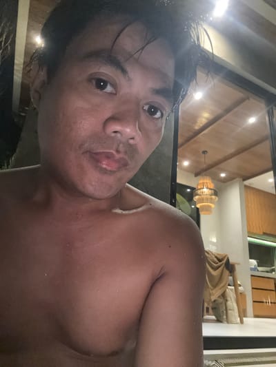 The image shows a shirtless individual with wet hair in an interior setting. The person has dark hair and is looking directly at the camera. The background includes a wooden ceiling with recessed lighting and a chandelier. A partially visible interior space with furniture is seen behind the person. The picture appears to have been taken indoors, possibly in a residential location or a resort-style setting. The location appears to be Tumbak Bayuh, Indonesia.
