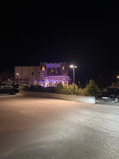 This nighttime photograph captures a modern architectural scene in Anatoliki Attiki, Greece. On the left, a multi-story building, possibly a hotel or residential complex, stands illuminated with warm interior lights visible through its windows. The building's facade features textured elements and is accented with vibrant purple lighting that highlights its structure. In front of the building, a large, curved pergola or canopy structure is also illuminated by purple lights, suggesting an outdoor lounge or dining area.

Lush green bushes and trees are strategically planted around the building and along the edge of a wide paved area, which dominates the right side of the frame and appears to be a parking lot or a spacious walkway. Modern streetlights with bright white lamps cast illumination onto the ground and parts of the architecture. A dark-colored car is parked in the upper right, displaying a partially visible license plate "FPH 7074". Another vehicle is faintly visible at the bottom right. The sky is completely dark, confirming the image was taken at night. The combination of warm interior lighting, bright white streetlights, and striking purple accent lights creates a sophisticated and inviting contemporary ambiance. No people or specific activities are depicted in this static outdoor environment.