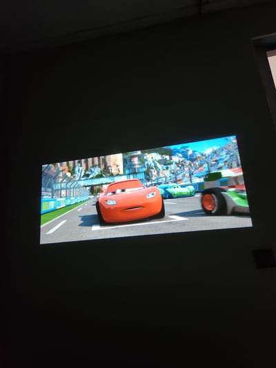 The image displays a projected scene of a cartoon race. In the foreground, a red race car, resembling Lightning McQueen from the movie Cars, is prominently featured. To its right, another race car with a green and white racing livery is partially visible, with its rear wheel turned towards the viewer. In the background, a green race car is seen on the track, and beyond that, a cityscape with buildings and a grandstand filled with spectators is depicted. The race appears to be taking place on a paved track with white lane markings. The environment suggests a race circuit within a city. There are no people or discernible interactions evident in the projected image. The overall scene is illuminated by the projection onto a dark surface, implying it is being viewed in a dimly lit room. The location is not specified within the image itself.
