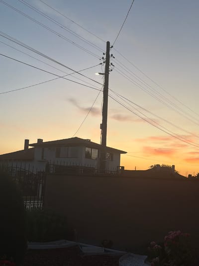 The image depicts a sunset scene over a residential area in Okorsh, Bulgaria. A wooden utility pole stands prominently in the center, supporting multiple power lines that crisscross the sky. The sky is illuminated by warm hues of orange and yellow, with scattered clouds tinged pink. In the background, a two-story house with a red tile roof is visible, featuring windows and chimneys. A dark, solid fence runs across the foreground, partially obscuring a wrought-iron fence and some greenery, including flowering bushes. The overall impression is of a quiet, suburban evening in a Bulgarian village. There are no people or animals visible in the frame.