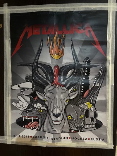 The image displays a poster for a Metallica concert. The poster features a graphic design with a central image of a goat's head with large horns, a melting candle on its head, and a black panther and a skull with yellow eyes emerging from below. The Metallica logo is prominently displayed at the top. Below the graphic, text indicates the date "21.7.2019" and the venue "LUZHNIKI STADIUM MOSCOW RUSSIA." The poster is taped to a dark surface, possibly a wall or door, with visible strips of masking tape along its edges. The lighting suggests it is an indoor setting. There are no people or actions depicted in the image, other than the static artwork of the poster. The location is identified as Luzhniki Stadium in Moscow, Russia, not Podkletnoe.