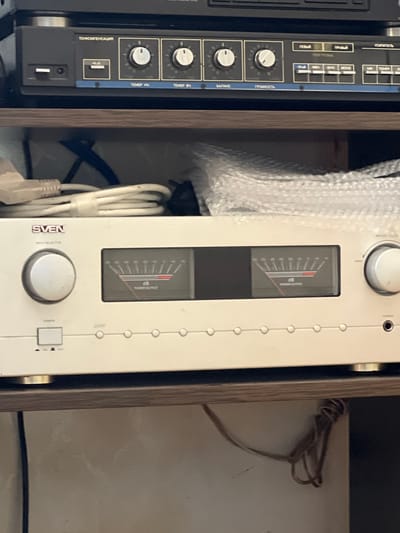 Two audio electronic components are closely positioned side-by-side within a wooden cabinet or shelf, viewed from a slightly elevated angle. The device on the left is darker, possibly black or dark grey, and features a classic design with numerous silver knobs and rectangular switches. Its front panel includes various controls for an amplifier, with labels in Cyrillic. Notable text includes "УСИЛИТЕЛЬ" (Amplifier), "ГРОМКОСТЬ" (Volume), "БАЛАНС" (Balance), "ТЕМБР НЧ" (Bass Tone), "ТЕМБР ВЧ" (Treble Tone), "ТОНКОМПЕНСАЦИЯ" (Loudness compensation), "ЛЕВЫЙ" (Left), "ПРАВЫЙ" (Right), "ПЕРЕГРУЗКА" (Overload), "МОНО" (Mono), and "ТЮНЕР" (Tuner).

To its right, a lighter, off-white audio component is visible. This device has a more streamlined design, dominated by two large, vertically oriented analog meters that display "dB" and "POWER OUTPUT". A prominent central knob is labeled "INPUT SELECTOR", and a power switch with "ON" and "OFF" indicators is located at the bottom right. The brand name "SVEN" is visible near the input selector knob. Part of a "PHONES" jack can be seen at the top right.

Bubble wrap rests on top of the lighter device, suggesting it might be recently acquired or stored. White coiled cables are tucked between the two components, while a darker, tangled cable is seen in the background to the right. The overall setting appears to be an indoor storage space, possibly a home, in Krayevsko-Armyanskoye, Russia.