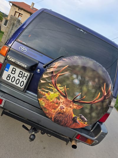 This is a close-up, angled shot of the rear of a dark blue Toyota Land Cruiser parked on a paved road. A spare tire cover featuring a graphic of a red deer with large antlers and two birds is mounted on the back. The license plate, which reads "BG B BH 8000", is visible below the "LAND CRUISER" emblem. To the left of the vehicle, a concrete fence and a two-story house with a red tiled roof are partially visible, indicative of a rural or suburban setting. The sky above is overcast with a few thin clouds. The image was captured in Suvorovo, Bulgaria, suggested by the Bulgarian license plate and the architectural style of the building. The scene appears to be a static display of the vehicle in its environment.