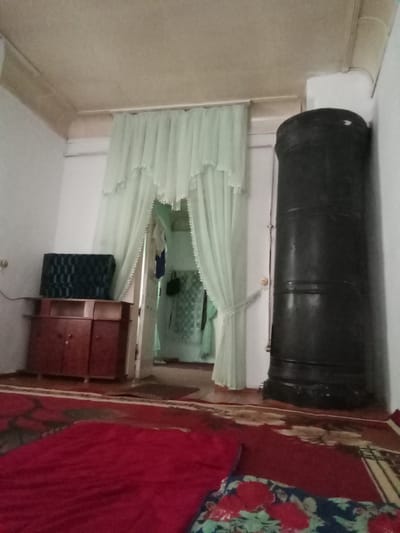 The image depicts an interior scene, likely a residential room, with a distinctive black, cylindrical stove prominently positioned on the right side. The stove has a textured surface with horizontal bands. To the left of the stove, sheer, light green curtains with decorative edging are drawn across an opening, revealing a glimpse of another room beyond. In the foreground, a portion of a red rug is visible, layered with a floral-patterned fabric. To the left of the curtains, a wooden cabinet with two doors and open shelves sits beneath a dark, patterned object, possibly a television or a speaker. The walls are painted white, and the ceiling is a neutral color. The lighting is dim, creating a subdued atmosphere. There are no people visible in the image. The setting appears to be a simple, functional living space. The mention of Karagana, Uzbekistan, suggests a geographical context for this domestic interior.
