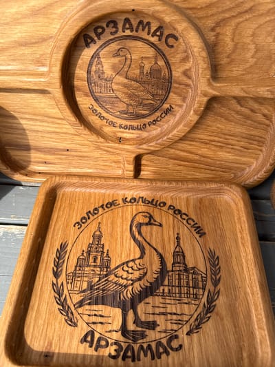 Two wooden serving platters are displayed side-by-side. Each platter is adorned with a circular emblem featuring an illustration of a swan and a church-like building. The word "Арзамас" (Arzamas) is inscribed on the left side of the circle, and "Золотое Кольцо России" (Golden Ring of Russia) is on the right. The platters are made of light brown wood. The rectangular platter has a border around its edges, while the other platter has a circular design with several indented compartments. The background features a wooden surface with a gray tone.